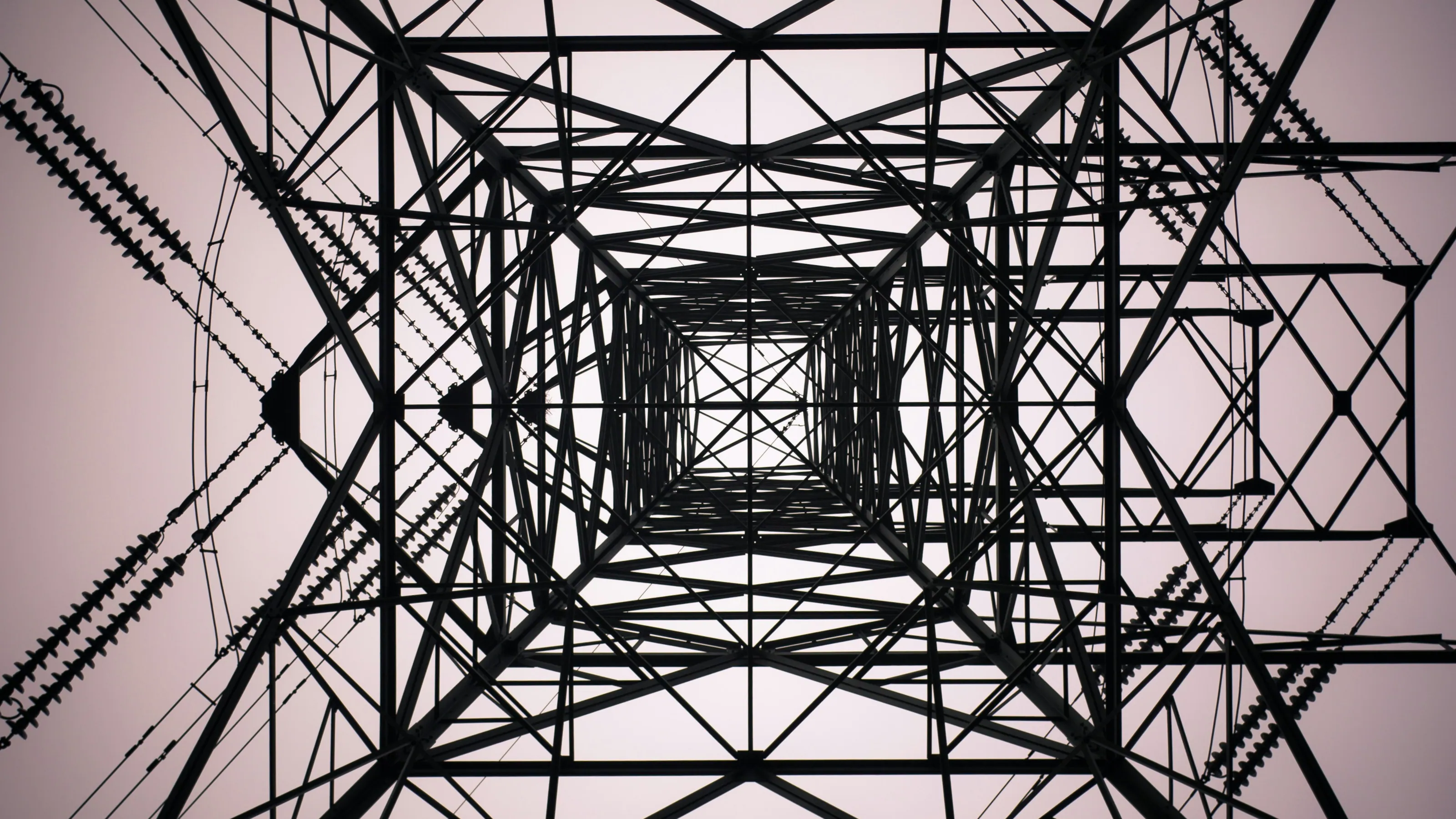 Looking straight up through the steel lattice of a high-voltage transmission tower, with insulator strings on the cross-arms