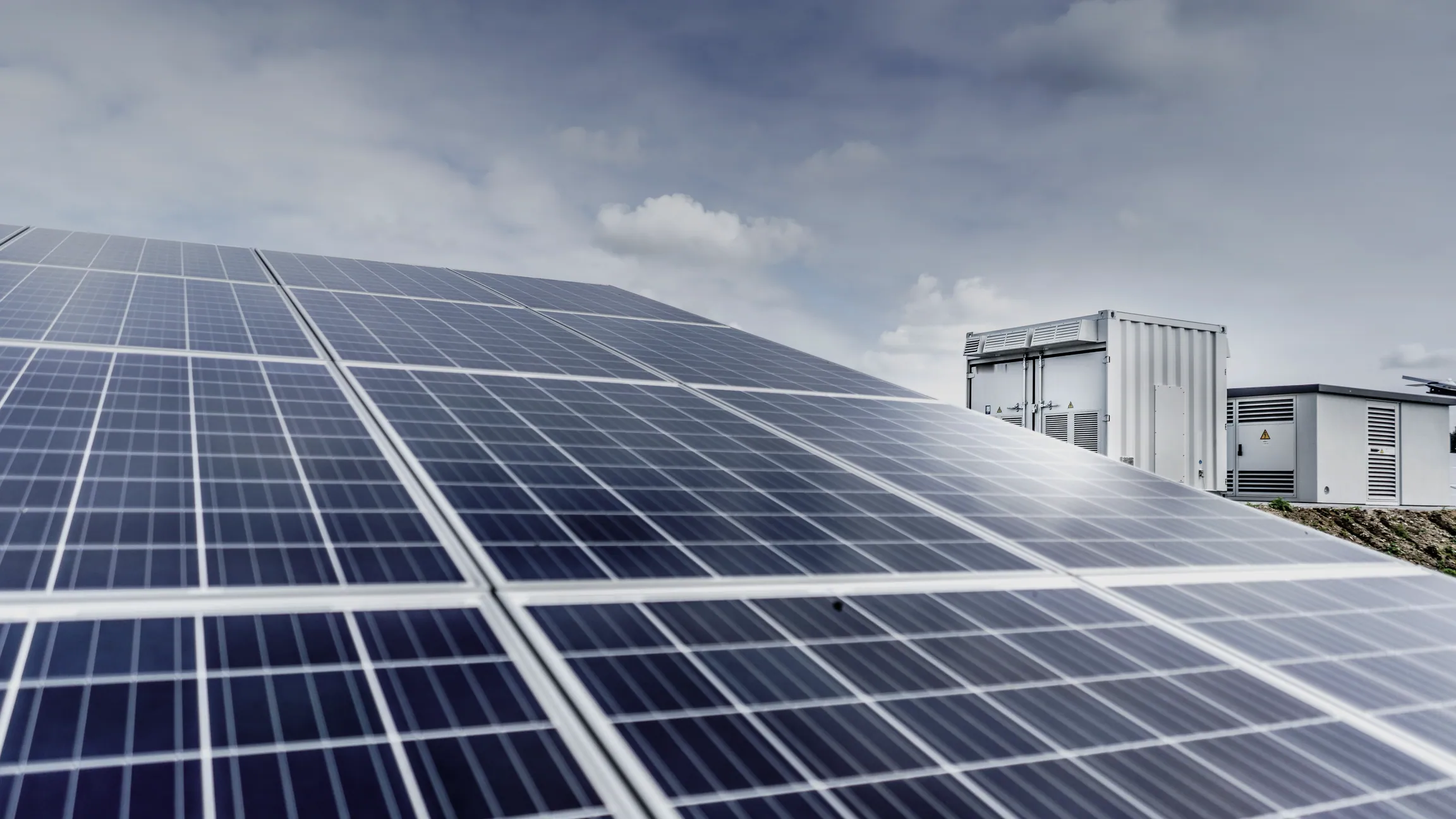 Ground-mounted solar panels in the foreground with battery storage and inverter containers behind them under a cloudy sky